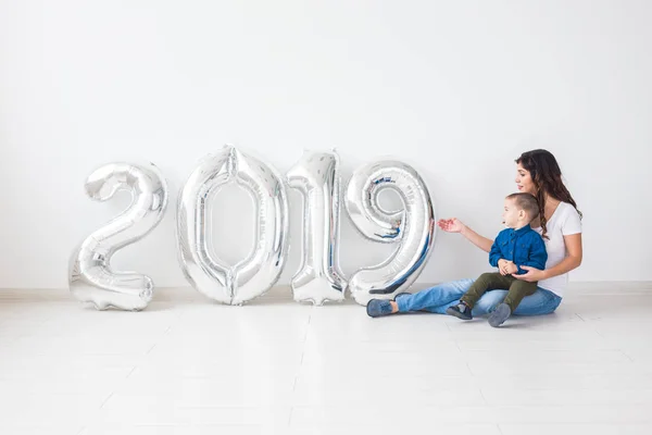 Año nuevo, celebración y concepto de vacaciones - madre e hijo sentados cerca de signo 2019 de globos de plata para el año nuevo en el fondo de la habitación blanca — Foto de Stock