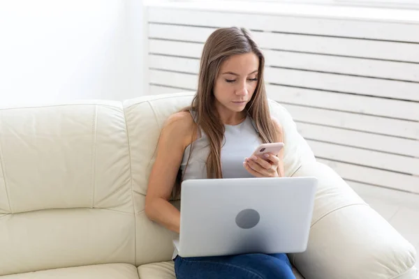 Teknik-, fritids- och människor koncept - ung kvinna sitter o vita soffan och chatta i netbook och mobile — Stockfoto