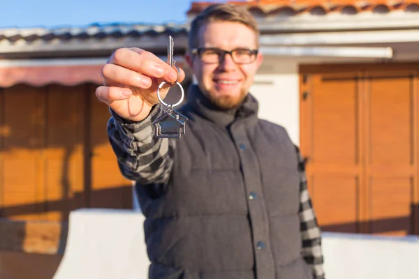 Happy house owner. Smiling young man holding keys and looking at camera while standing against new home background — Stock Photo, Image