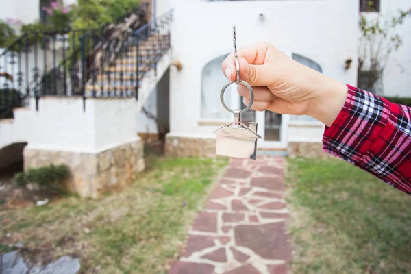 Concepto de bienes raíces y bienes raíces - La mano está sosteniendo las llaves de la casa en forma de llavero frente a una nueva casa —  Fotos de Stock