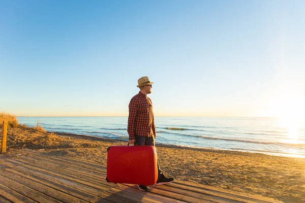 Concepto de viaje, viaje, verano y vacaciones: un hombre con un estuche rojo está parado en una playa y viendo una puesta de sol — Foto de Stock