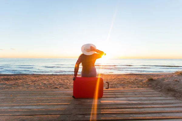 Viaggio, vacanza e concetto di persone - turista donna seduta vicino al mare sulla valigia rossa e in cappello e guardando il tramonto — Foto Stock