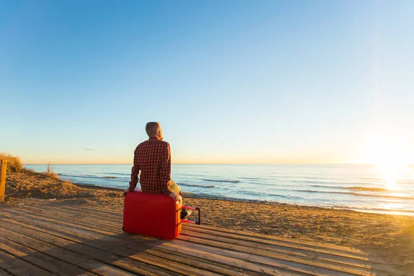 Vacanze, viaggi e vacanze estive concetto un uomo seduto sulla valigia rossa e guardando il tramonto — Foto Stock