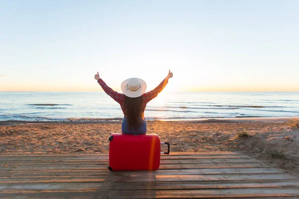 Viaggiare, vacanze e persone concetto - donna felice seduta su una valigia rossa in un cappello di fronte a voi, guardando il mare — Foto Stock