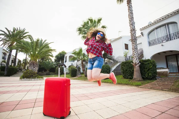 Путешествия, туризм, эмоции и концепция людей - счастливая молодая женщина прыгнула в шляпу и солнечные очки рядом со своим красным чемоданом — стоковое фото