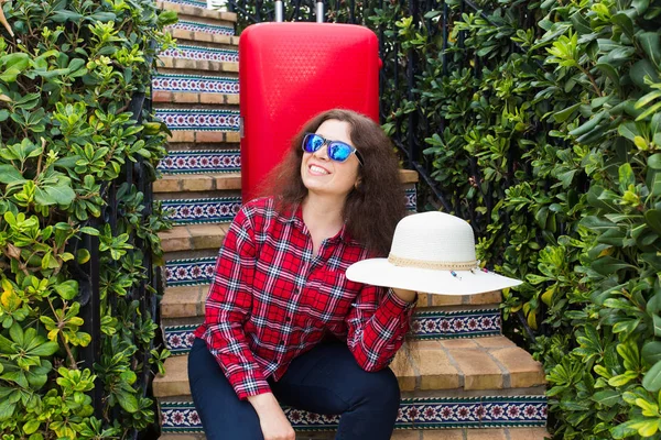 Reizen, vakanties en feestdagen concept - gelukkig reiziger vrouw zittend op trap in zonnige bril met rode koffer en hoed — Stockfoto