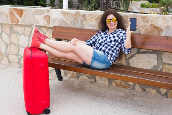 Путешествия, туризм, технологии и человеческая концепция - счастливая молодая женщина сидит на скамейке, ставит ноги на чемодан и показывает свой телефон — стоковое фото