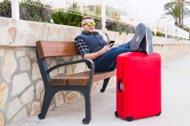 Holidays, travel and rest concept - portrait of young man sitting on bench outdoors with legs on suitcase