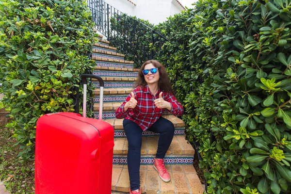 Концепція відпочинку, подорожей та туризму - молода жінка з червоною валізою, що сидить на сходах і показує великі пальці вгору — стокове фото