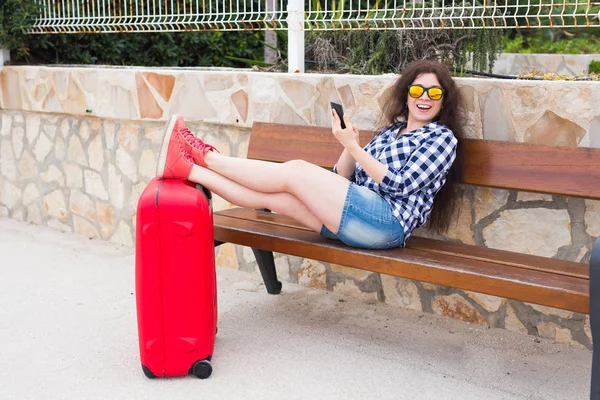 Технология и концепция путешествия - молодая женщина со смартфоном и чемоданом сидит на скамейке запасных — стоковое фото