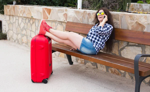 Travel, tourism, technology and people concept - happy young woman sit on bench and put her feet on suitcase and talking on mobile. She is ready to travel