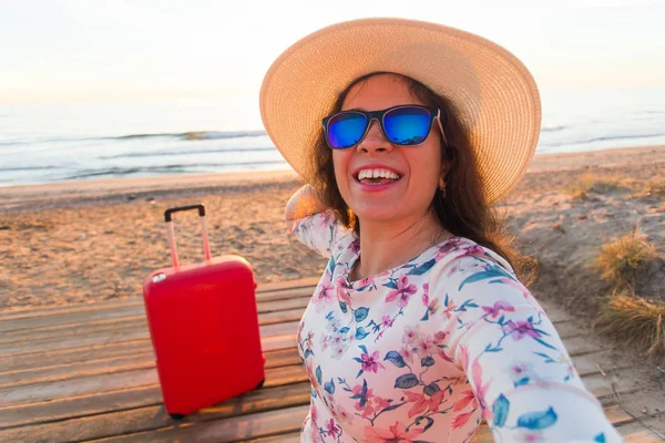 Подорожі, свято і концепція людей - молода жінка в капелюсі і окулярах стоїть на пляжі з червоною валізою і посміхається — стокове фото