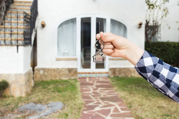 Real estate and property concept - Hand is holding house keys on house shaped keychain in front of a new home — Stock Photo, Image
