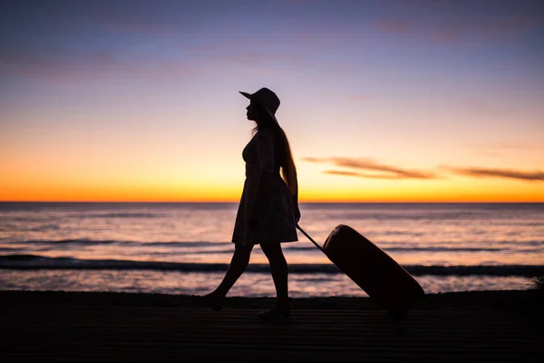 Relax woman with suitcase on a beach at sunset silhouette. Holiday travel concept. Young lady with suitcase on ocean landscape background