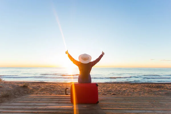 Viajes, vacaciones y concepto de personas - turista sentada cerca del mar en la maleta roja y en el sombrero y viendo la puesta de sol y mostrar los pulgares hacia arriba — Foto de Stock