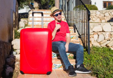 İnsanlar, tatil, seyahat ve dinlenme kavramı - merdivenlerde oturan ve gösterilen genç adam bavulunu yaşasın