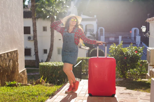 Viajes, turismo y el concepto de la gente - mujer joven feliz en el sombrero con una enorme maleta roja de pie cerca de las casas y sonrisa — Foto de Stock