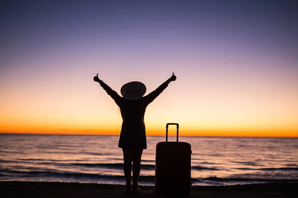 Viajes, vacaciones y concepto de vacaciones - Mujer joven de pie con maleta en la playa de arena en el mar al atardecer — Foto de Stock