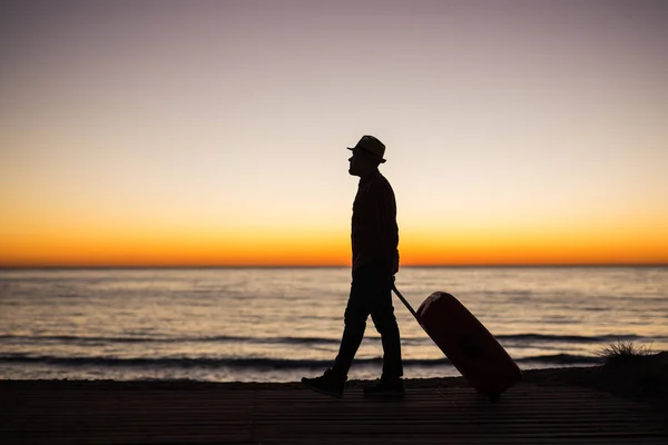 Concepto de vacaciones, verano y viajes: silueta de hombre joven con maleta al atardecer cerca del mar — Foto de Stock