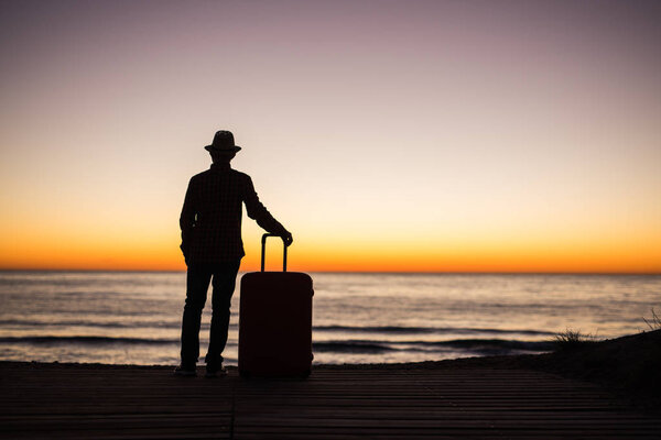 Relax man with suitcase on a beach at sunset silhouette. Holiday travel concept. Guy with suitcase on ocean landscape background
