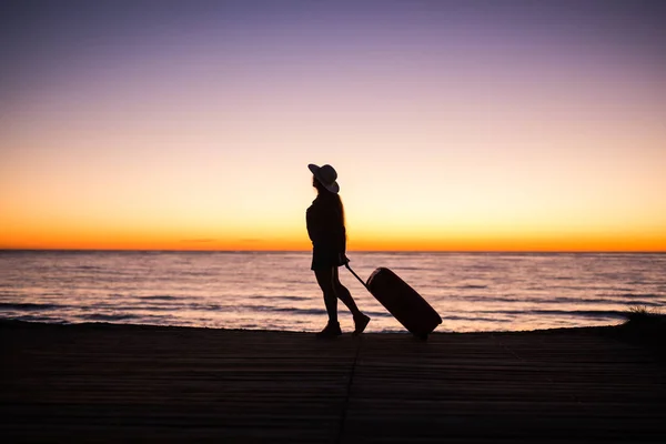 Viajar, playa y concepto de vacaciones - silueta femenina caminando a lo largo de la costa del océano tirando de la maleta — Foto de Stock