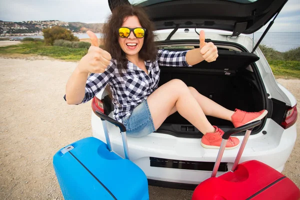 Mladá žena sedí v zadní části vozu s úsměvem a ukazuje palec. Dívka se smíchem v otevřeného kufru auta. Letní výlet — Stock fotografie