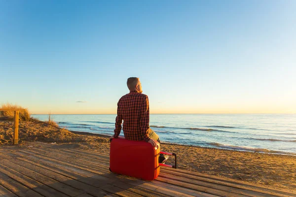 Vacaciones, viajes y vacaciones de verano concepto de un hombre sentado en la maleta roja y ver la puesta de sol — Foto de Stock