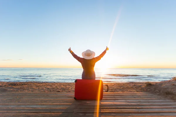 Viajar, vacaciones y concepto de personas - mujer feliz sentada en una maleta roja en un sombrero frente a usted, mirando el mar — Foto de Stock