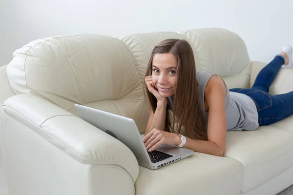 Технологии, фрилансер и концепция людей - красивая молодая женщина лежит на диване с ноутбуком и смотрит в камеру — стоковое фото