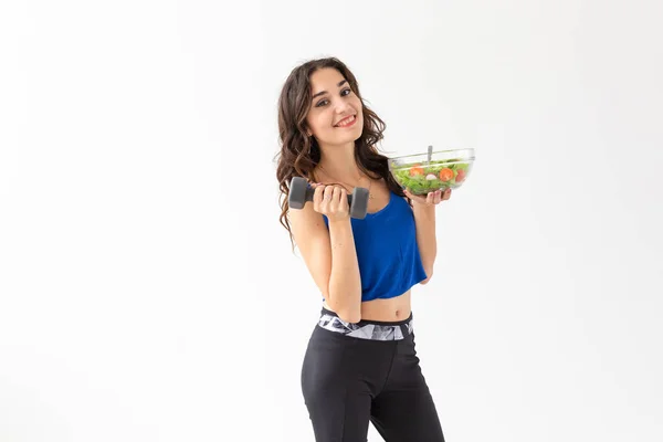 Stile di vita sano, dieta, fitness e concetto di persone: la giovane donna sta mangiando un'insalata e facendo sport con manubri su sfondo bianco — Foto Stock