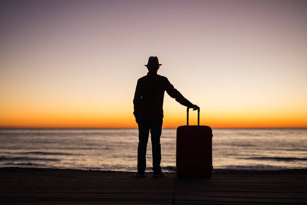 Vacation, summer and travel concept - young man silhouette with suitcase at sunset near the sea