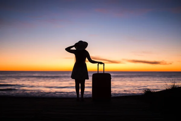 Viajes, vacaciones y concepto de vacaciones - Mujer joven de pie con maleta en la playa de arena en el mar al atardecer — Foto de Stock