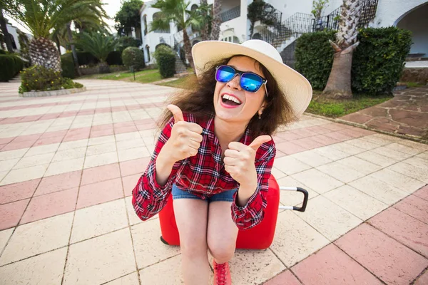 Reizen, toerisme en vakantie concept - grappige jonge vrouw zittend op rood koffer in de bril en muts duim opdagen en glimlachen — Stockfoto