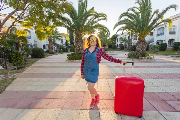 Reizen, toerisme en mensen concept - gelukkige jonge vrouw in een hoed met een enorme rode koffer staande in de buurt van huizen en glimlach — Stockfoto