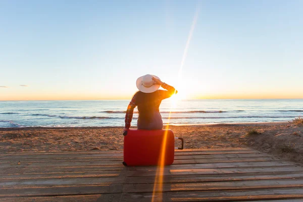 Viaggio, vacanza e concetto di persone - turista donna seduta vicino al mare sulla valigia rossa e in cappello e guardando il tramonto — Foto Stock