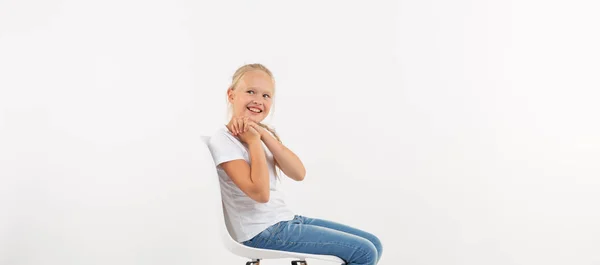 Alegre niña rubia pequeña sentada en la silla con sonrisa aislada sobre fondo blanco con espacio para copiar — Foto de Stock