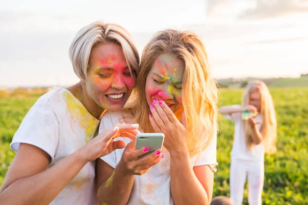 Indiase festival van holi, mensen concept - twee lachende meisjes met kleurrijke poeder op de gezichten kijken naar de smartphone — Stockfoto