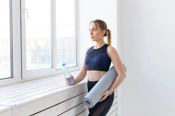 Gente, deporte y fitness concepto - mujer joven con esterilla de yoga y botella de agua de pie cerca de la ventana — Foto de Stock