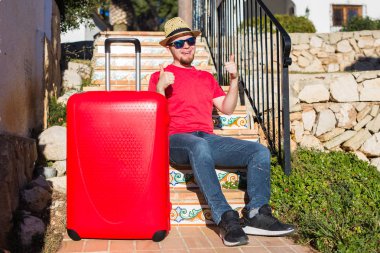 İnsanlar, tatil, seyahat ve dinlenme kavramı - merdivenlerde oturan ve gösterilen genç adam bavulunu yaşasın