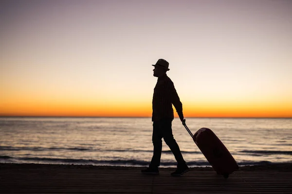 Relájese hombre con la maleta en una playa al atardecer silueta. Concepto de viaje de vacaciones. Tipo con maleta en el fondo del paisaje oceánico — Foto de Stock
