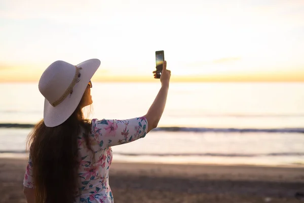Technology, travel and holidays concept - Young woman takes pictures on the smartphone sunrise at the sea