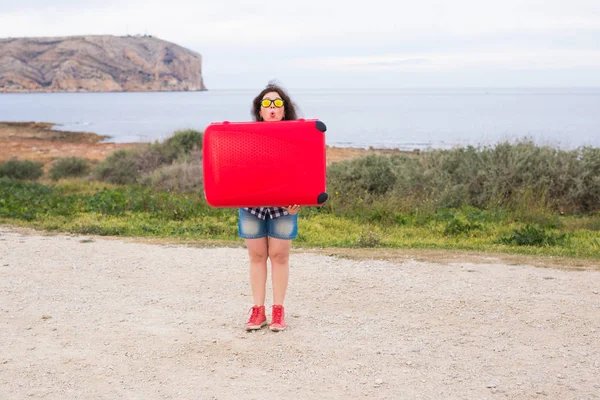 Concepto de viaje, turismo y gente: una chica feliz sosteniendo una enorme maleta roja sobre su cabeza, con gafas divertidas y sonriendo en el fondo de la naturaleza. — Foto de Stock