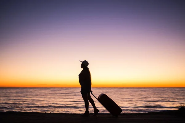 Viajar, playa y concepto de vacaciones - silueta femenina caminando a lo largo de la costa del océano tirando de la maleta — Foto de Stock