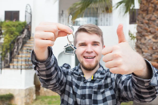 Happy house owner. Smiling young man holding keys and looking at camera while standing against new home background — Stock Photo, Image