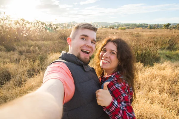 Koncepcja podróży, turystyka i przyroda - uśmiecha się para biorąc selfie na pole i pokazuje kciuk w górę — Zdjęcie stockowe
