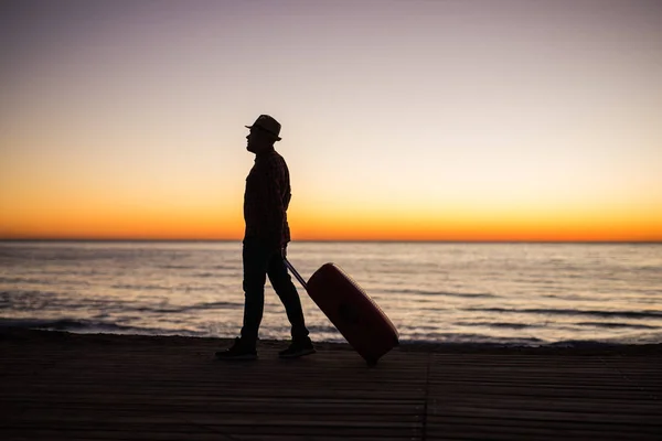 Concepto de vacaciones, verano y viajes: silueta de hombre joven con maleta al atardecer cerca del mar — Foto de Stock