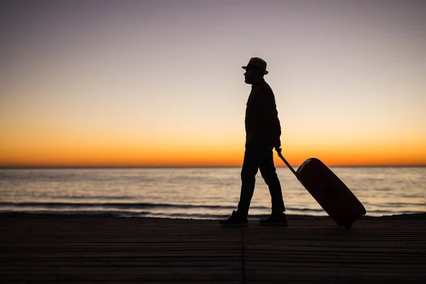 Concepto de vacaciones, vacaciones y viajes - Silueta de hombre joven con maleta en la playa al amanecer . — Foto de Stock