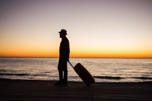 Concepto de vacaciones, verano y viajes: silueta de hombre joven con maleta al atardecer cerca del mar — Foto de Stock