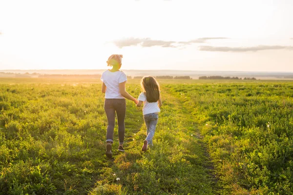 Vista trasera de la madre y la hija corriendo en campo verde con puesta de sol en el fondo —  Fotos de Stock