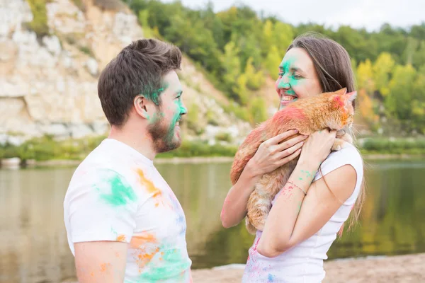 Festival holi, feriados, turismo, animal de estimação e conceito de natureza - Retrato de mulher e homem com gato coberto de poeira multicolorida — Fotografia de Stock